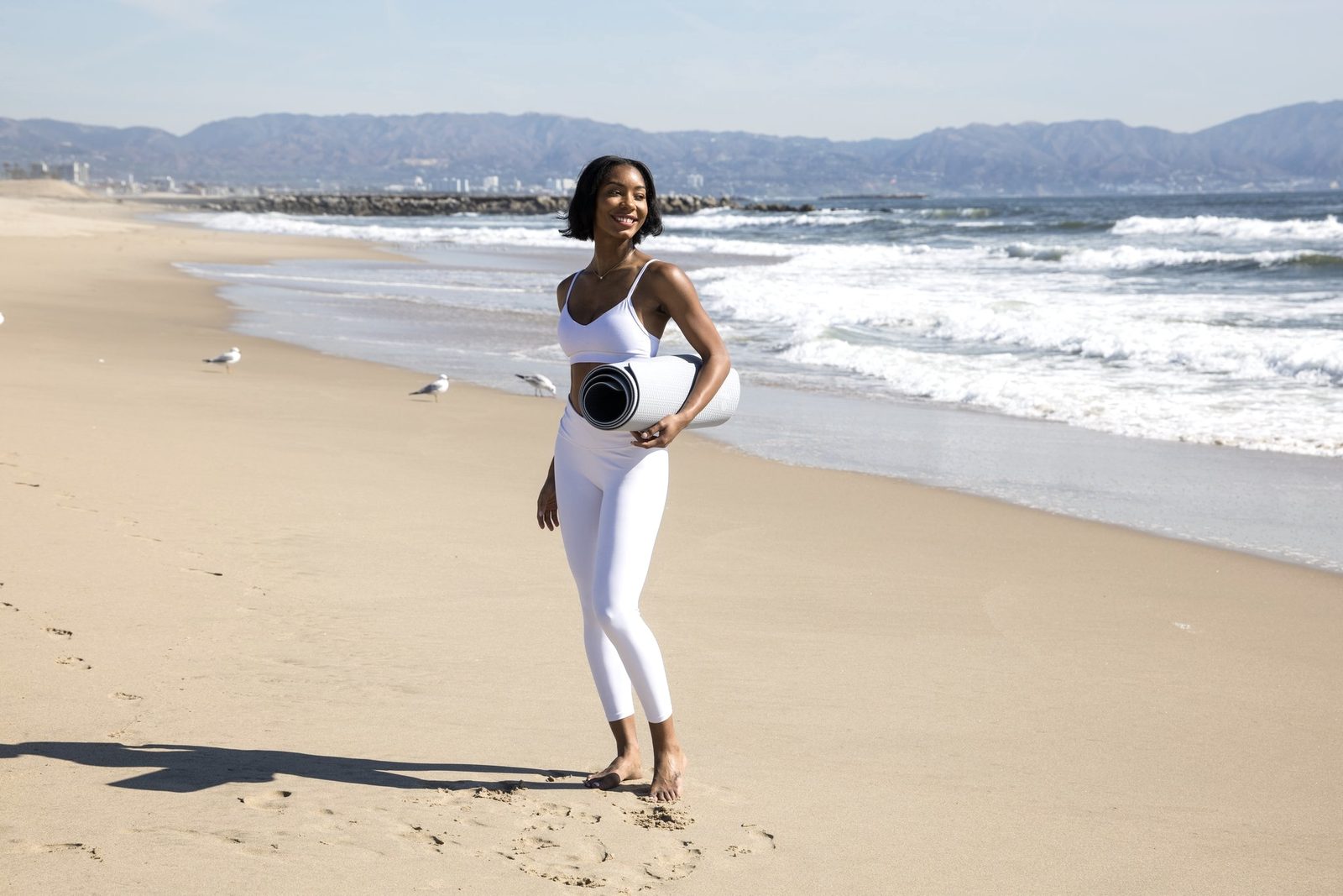 Maya smiling on the beach, holding her rolled yoga mat, the surf behind her
