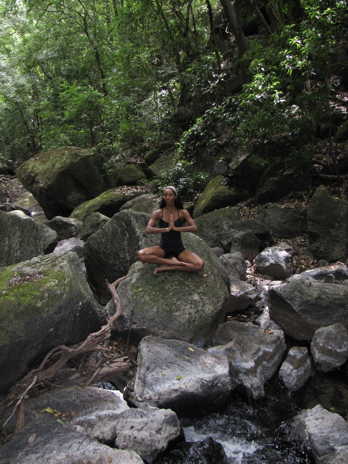 Maya seated in meditation on a mossy boulder above a forest stream, palms together