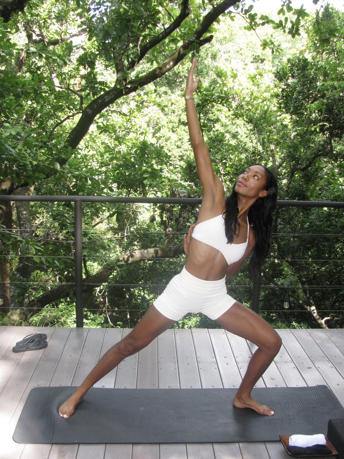 Maya reaching one arm skyward in a wide warrior pose on a treetop deck